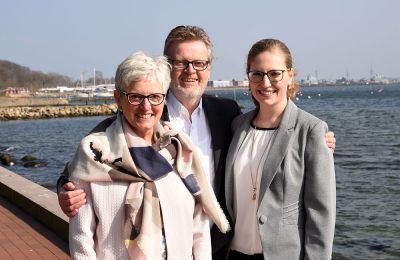Drei lächelnde Personen posieren im Business-Outfit am Wasser bei sonnigem Wetter.